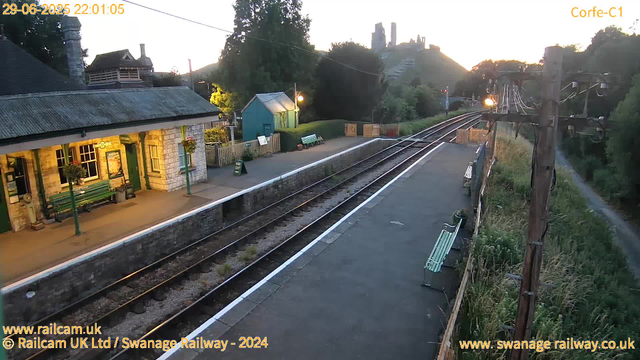 A small train station platform at sunset with a building on the left side, decorated with hanging flower baskets and benches. There are railway tracks running parallel to the platform, which is bordered by a low stone wall. A green bench is visible on the platform, along with a small green shed and some signage. Overhead, power lines and utility poles run alongside the tracks, and a path with tall grass is on the right side. In the background, there are hills or mountains with some buildings or ruins atop them, illuminated by the fading sunlight. The scene appears calm and quiet.
