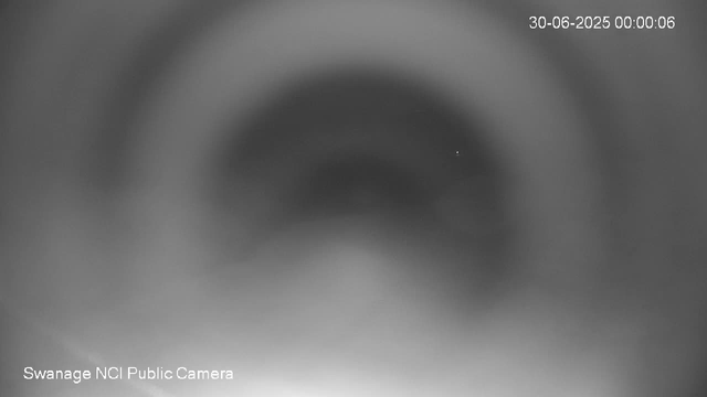 A blurry, grayscale close-up image from a webcam showing a dark circular area in the center with a small bright dot inside it. The top right corner displays a timestamp reading "30-06-2025 00:00:06," and the bottom left corner notes "Swanage NCI Public Camera."