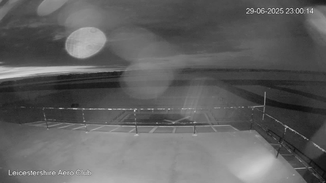 A black-and-white image captured by a webcam shows the tarmac and perimeter fence of Leicestershire Aero Club. The sky appears cloudy, and there are some bright spots that could be reflections or light artifacts. The timestamp indicates it was taken on June 29, 2025, at 11:00:14 PM.