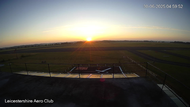 The image shows a sunrise at Leicestershire Aero Club with the sun beginning to rise over a flat, open landscape. The sky is mostly clear with a few wispy clouds. In the foreground, there is a helipad with a large black "H" painted on it, surrounded by a safety barrier. The area appears to be part of an airfield, with runways and grassy fields extending into the distance. A timestamp in the top right corner reads "30-06-2025 04:59:52".