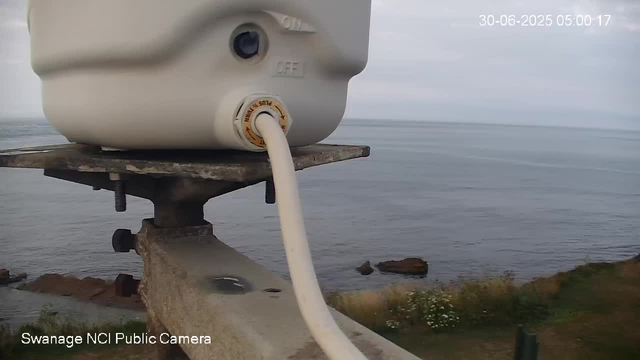 A white device is mounted on a metal platform overlooking a body of water with rocks near the shore. The device has a cable connected to it, and the background shows a calm ocean under a cloudy sky. The timestamp in the upper right corner indicates the date and time as June 30, 2025, at 5:00 AM.