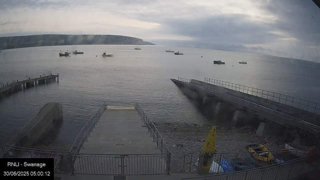 A view of a coastal harbor with several boats floating on calm water under a cloudy sky. A concrete pier extends into the water in the foreground, with a yellow caution sign and some equipment nearby. In the background, there are cliffs along the shoreline. The scene appears to be early morning.