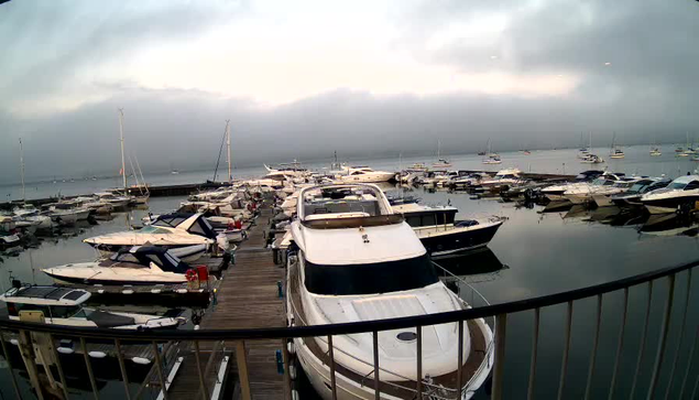 A marina filled with various boats and yachts docked along a wooden pier. The water is calm and reflects the boats. The sky above is overcast with dark clouds, suggesting an approaching storm or cloudy weather. In the foreground, a metal railing runs across the bottom of the image.
