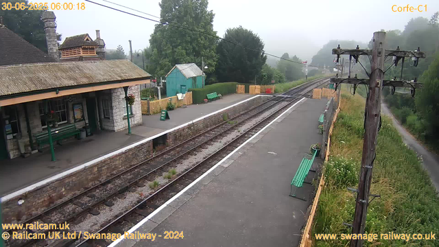 A small countryside train station platform with a stone building on the left featuring a tiled roof and a chimney. There are green benches on the platform, some with flowers hanging from the trash bin. A wooden fence surrounds a small green shed in the background. Overhead, empty railway tracks extend into a misty, tree-lined landscape. A tall wooden utility pole with insulators and wires stands on the right side of the platform. The scene appears foggy and quiet early in the morning.