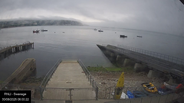 A harbor scene on a cloudy day with several boats anchored in calm water. There are docks on the left and right sides, with a concrete platform in the foreground. A yellow buoy and some equipment are near the bottom right corner. In the background, there are distant hills partially obscured by fog or low clouds. The timestamp indicates the image was captured early in the morning.