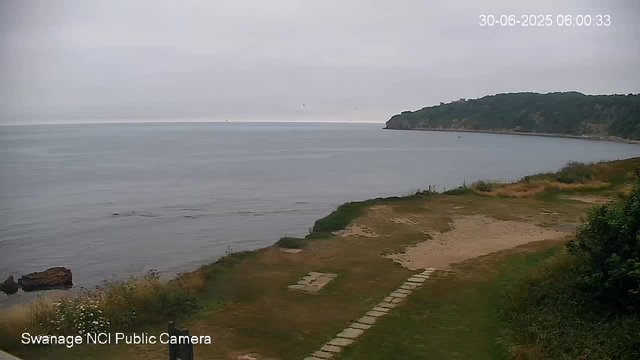 A view of a cloudy day at a beach with a narrow pathway leading down to the shore. The beach has a mix of sand and grassy patches, and there is a hill covered with trees in the background stretching along the coast. The water appears calm with gentle waves, and a few small rocks are visible near the shoreline. The timestamp in the top right corner indicates the date as June 30, 2025, and the time as 6:00 AM.