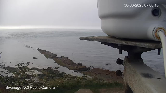A view from a webcam showing a body of water with a partially submerged, straight, stone breakwater extending into the sea. The sky is overcast and gray, and there is a large white object with a cable attached in the upper right corner of the image. The timestamp on the image reads "30-06-2025 07:00:13."