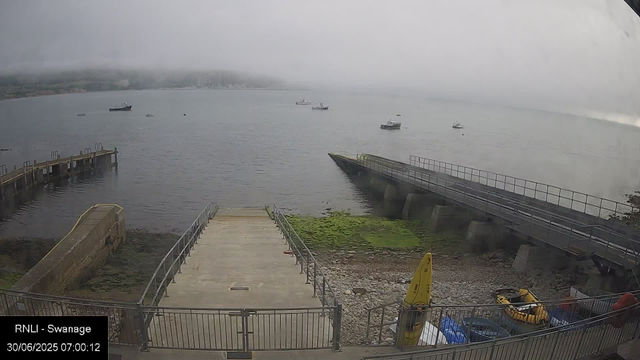 A view of a misty harbor with a concrete pier leading into the calm water. Several boats are anchored in the distance, with some near the shore and others farther out. There is a grassy patch and a yellow safety sign at the bottom right of the image, along with some equipment and containers. The sky is overcast with low-hanging clouds, and the overall scene has a cool, muted tone.