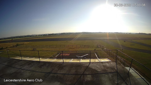 A view from a webcam at Leicestershie Aero Club shows a clear day with the sun shining brightly in the sky. The image captures an open grassy area with runways or taxiways in the background, and a section of a rooftop or balcony with a metal railing in the foreground. The time and date are displayed in the top right corner as 30-06-2025, 07:00:08.