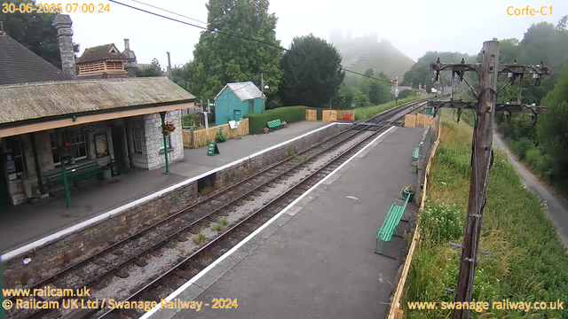 A small railway station platform with a stone building on the left, featuring a corrugated roof and hanging flower baskets. There are green benches and a green "Way Out" sign on the platform. Parallel train tracks run through the scene, with some small plants growing between the sleepers. To the right, there is an old wooden utility pole with multiple wires and a narrow dirt path running alongside the platform into a green, tree-filled landscape. The background shows a foggy view of distant hills with some structures barely visible through the mist. The image has a timestamp of 30-06-2025 07:00:24 and is labeled with "Corfe-C1".