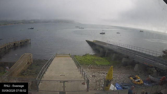 A view of a harbor with several boats floating on calm water, a concrete dock extending into the water, a grassy and rocky shoreline, and a cloudy, misty sky overhead. There are a few yellow safety equipment items and people visible near the shore. The bottom left corner shows a timestamp and location: RNLI - Swanage, 30/06/2025 07:59:52.