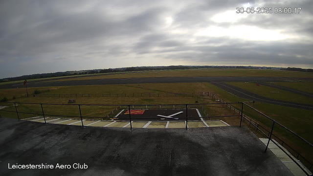 A view from a webcam at Leicestershire Aero Club showing a cloudy sky, an open grassy area, a small runway with painted markings, and a section of a helicopter landing pad with a white 'X' on a black background. The date and time are displayed in the top right corner as "30-06-2025 08:00:17," and the bottom left corner contains the text "Leicestershire Aero Club."