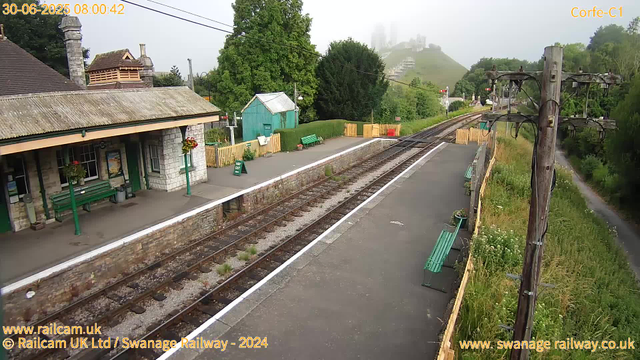 A small railway station platform with a stone building on the left, featuring green benches and hanging flower baskets. There are potted plants and a "WAY OUT" sign on the platform. The railway tracks run through the center of the image, with a yellow fence on the far side. To the right, wooden utility poles with wires run alongside a grassy area with wildflowers. In the background, lush green trees and a foggy hillside with buildings or castle ruins atop are visible. The scene is overcast and hazy. The timestamp indicates the photo was taken at 8:00 AM on June 30, 2025.