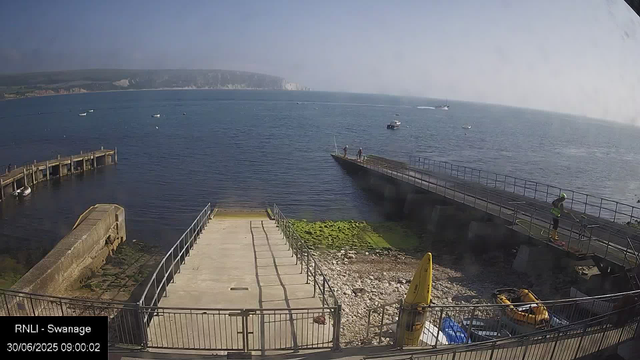 A view of a coastal dock area with a concrete pier extending into the water. Two people are standing at the end of a small boat on the pier, and a person in a high-visibility vest and helmet is walking along the ramp on the right. There are a few boats anchored in the water, and a small inflatable boat is on the ground near a yellow kayak. The background features the open sea with several boats and a distant shoreline with white cliffs or buildings. The scene is bright with clear skies.