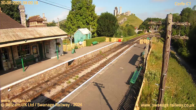 A railway station platform with a building on the left side, featuring stone walls, a tiled roof, and hanging flower baskets. There are benches on the platform and a green sign reading "WAY OUT." A small green shed is visible further along the platform, next to a wooden fence separating the station from the railway tracks. The tracks run from the bottom left toward the horizon, with overhead electrical lines supported by wooden poles. To the right of the tracks, a narrow canal with rippling water runs parallel, bordered by wild grass and bushes. In the background, a hill with a castle ruins perched on top, surrounded by green trees, is visible under a clear blue sky. The timestamp on the image reads "30-06-2025 09:00:18."