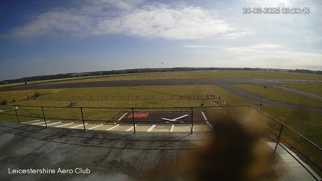 An empty rectangular helipad with a white "H" in the center is visible, surrounded by a black fence. Beyond the helipad, there is a grassy area, and further in the distance is a runway with a small airplane taking off or landing. The scene is under a partly cloudy sky with patches of sunlight. The timestamp in the upper right corner reads "30-06-2025 09:00:43," and the bottom left corner has the text "Leicestershire Aero Club."