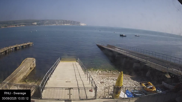 A view of a calm seaside scene with a concrete pier extending into the water, a yellow caution sign, and rescue equipment with boats visible nearby. In the distance, a gentle hillside with white cliffs lines the horizon under a partly cloudy sky.