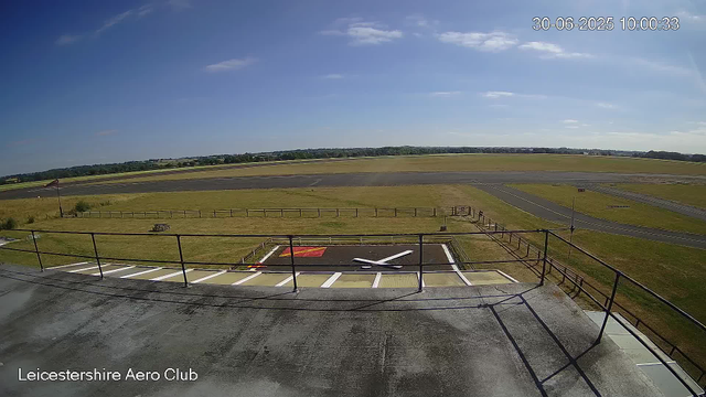 An empty aircraft hangar with a helipad marked by a white cross on the ground. The hanger is surrounded by a black metal railing and is located on a paved area that extends outward. Beyond the hangar, there is a grassy field with a picnic table, and further out, an airport runway stretching across the horizon. The sky above is bright and partly cloudy. The top right corner shows the date and time: 30-06-2025, 10:00:33. The bottom left corner has the text "Leicestershire Aero Club."