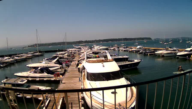 A marina filled with various boats and yachts docked along a wooden pier, with calm water and a distant shoreline under a clear, blue sky.