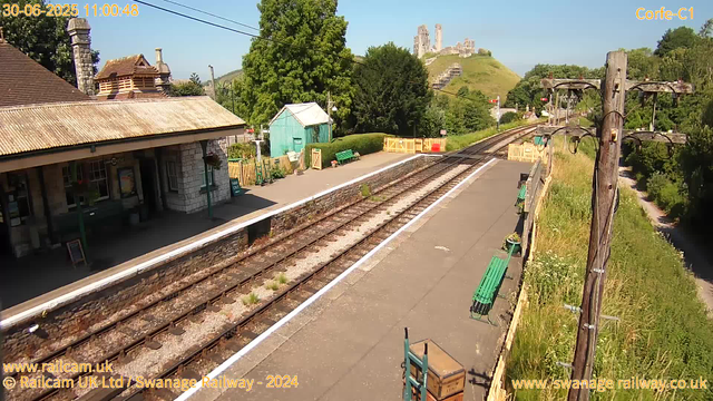 A small train station platform with railway tracks running through it. There is a building with a chimney and tiled roof on the left side, and a small green shed with a white roof in the background. Several green benches are situated along the platform, and there are wooden utility poles with wires running overhead on the right side. In the distance, a castle is visible on a hill, surrounded by green trees and fields. The sky is clear and blue, and it appears to be a sunny day. The timestamp indicates the date as June 30, 2025, at 11:00:48.