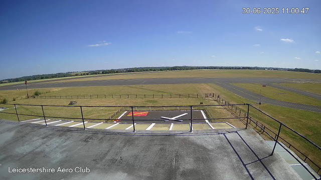 A view from a webcam at Leicestershire Aero Club shows a clear daytime sky with a few small clouds. In the foreground, there is a helipad marked with a large white "H" and surrounding painted lines, with a red and yellow safety area nearby. The helipad is enclosed by a black metal fence and has a railing on a balcony or observation platform from which the photo is taken. Beyond the helipad, there is a grassy area with a picnic table, a small bush, and a flagpole. In the background, an airstrip with multiple taxiways and runways stretches towards the horizon, with trees and fields in the distance. The timestamp on the image reads "30-06-2025 11:00:47," and the location is labeled "Leicestershire Aero Club" in the bottom left corner.