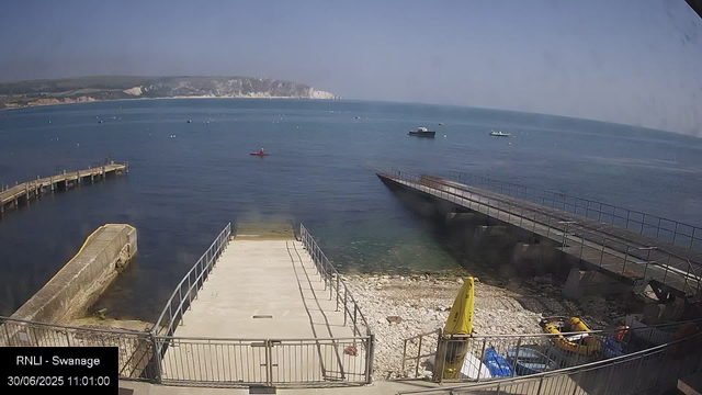 A view of a seaside dock area with calm water, a breakwater on the left, and a ramp leading into the water. Several boats are floating in the distance, and there are cliffs or hills along the coast visible at the horizon. The weather appears clear and sunny.