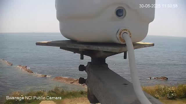 A close-up view of a white device with a cable plugged into it, mounted on a metal bracket. In the background, there is a scenic view of the ocean with some rocky formations jutting into the water and a grassy shoreline with yellow and white flowers. The sky is clear and bright. The image includes a timestamp in the upper right corner and a label indicating it was captured by a "Swanage NCI Public Camera."
