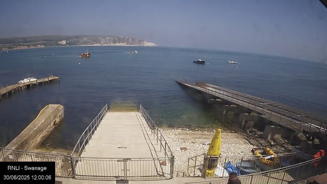 A view of a harbor with a concrete pier extending into calm blue water, boats floating in the distance, and white chalk cliffs in the background. There is a yellow caution sign, a small yellow inflatable boat, and some equipment near the shore, with a person partially visible at the bottom of the image. The scene is captured during the daytime with clear weather.