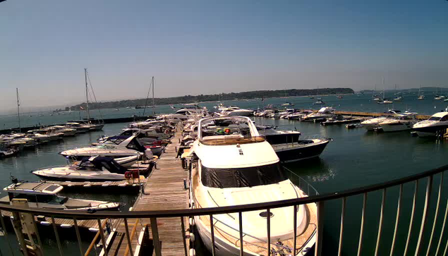 A wooden dock extends into a harbor filled with various boats and yachts, with additional boats anchored farther out on the calm water. The sky is clear and blue, and a distant shoreline is visible on the horizon. The perspective is from behind a curved metal railing at the edge of the dock.