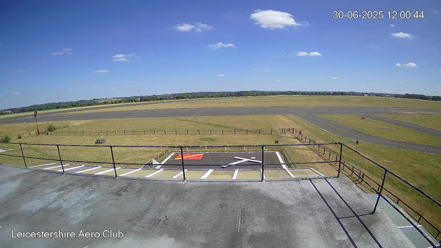 An empty airport runway and taxiway viewed from a high vantage point on a clear, sunny day. There is a grassy area with a small fenced section and a few signs near the foreground. The sky is mostly clear with a few scattered clouds, and the horizon shows distant trees and land. The timestamp on the image reads "30-06-2025 12:00:44" and the location is labeled "Leicestershire Aero Club."