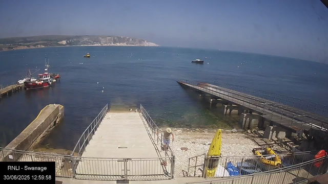 A view of a harbor with a concrete pier extending into calm blue water. Several boats are docked, and a few are out at sea. There are a few seagulls perched on the pier and floating in the water. In the foreground, a person wearing a wide-brimmed hat is walking along the lower part of the harbor. On the right, there is some rescue equipment, including a yellow life raft and buoys, and a yellow caution sign. The background shows a distant coastline with white cliffs under a clear, slightly hazy sky. The timestamp indicates the image was taken on June 30, 2025, at 12:59 pm, at the RNLI (Royal National Lifeboat Institution) station in Swansea.