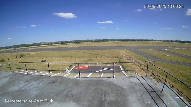 A wide view of an airfield on a sunny day with a clear blue sky and scattered white clouds. In the foreground, there is a concrete balcony with a metal railing, and a helicopter pad marked with a red square and a white helicopter symbol. The grassy area surrounds the tarmac, which has taxiways and runways stretching into the distance. A few small structures and signs are visible near the runways. The date and time displayed in the top right corner are June 30, 2025, at 13:00:24. The bottom left corner has the text "Leicestershire Aero Club."