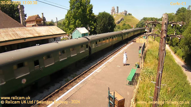 A vintage green train is stopped at a small countryside station on a sunny day. A woman dressed in white is standing on the platform, looking at the train. There are a few benches and a wooden luggage cart on the platform. To the right, there are tall wooden utility poles with wires extending into the distance. In the background, lush green trees surround the area, and a hillside with ruins of an ancient castle or fortress is visible under a clear blue sky. The date and time in the corner indicate June 30, 2025, at 1:00 PM.