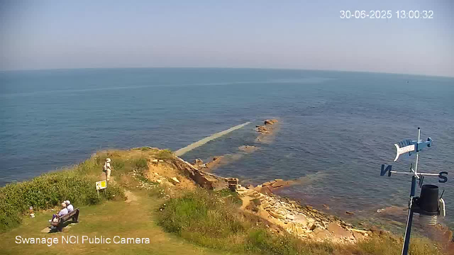 A scenic view of a seaside location with a grassy area and a stone pier extending into the calm ocean. Two people are sitting on a bench in the grass, and another person is standing near some bushes, looking out towards the water. The sky is clear, and the timestamp shows the date as June 30, 2025, at 1:00 PM. There is a weather vane on the right side of the image. The camera is labeled "Swanage NCI Public Camera."