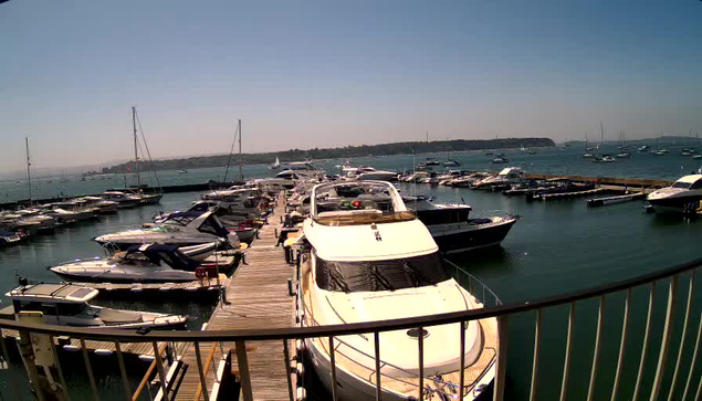 A wide view of a marina filled with various boats and yachts docked along wooden piers. The scene is set on a sunny day with a clear blue sky, and there are multiple sailboats and motorboats on the water. A black metal railing is visible in the foreground, curving along the edge of the viewpoint. In the background, a distant shoreline with some structures and boats can be seen.