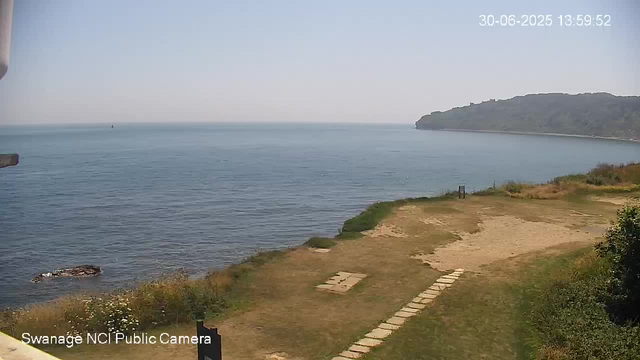 A view of a peaceful coastal scene on a clear day, showing calm blue water meeting the sky at the horizon. To the right, there is a tree-covered headland extending into the water. In the foreground, a grassy area with a stone path leads toward the water, with a small bench nearby. The image is timestamped "30-06-2025 13:59:52" in the upper right corner, and "Swanage NCI Public Camera" is written in the bottom left corner.