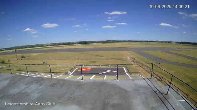 An outdoor view of a clear, sunny day at Leicestershire Aero Club, showing an airfield with runways and grassy areas. The sky is blue with scattered small white clouds. There is a small guardrail and a marked parking area in the foreground. In the background, open fields and a distant tree line are visible. The timestamp in the top right corner reads "30-06-2025 14:00:21."