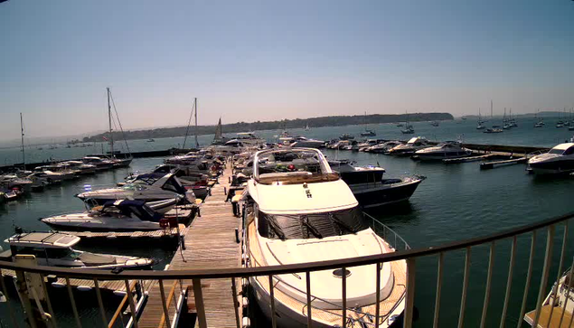 A dock with several yachts and boats moored on either side, extending into a harbor. The water appears calm, and there are additional boats and sailboats visible in the background. The sky is clear and blue.
