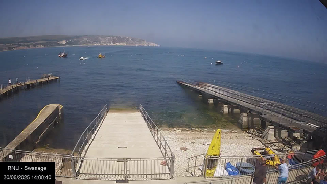 A coastal scene with a pier extending into calm blue water. Several boats are anchored offshore, and a distant shoreline with cliffs is visible under a clear sky. In the foreground, there are people near the water, some standing and chatting, along with equipment like a yellow kayak and life jackets. The image is labeled “RNLI - Swanage” with the date and time “30/06/2025 14:00:40”.
