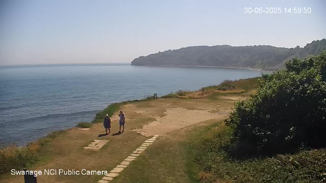A coastal scene with a sandy and grassy path leading to the water. Two people wearing hats and casual clothing are walking along the path. The calm sea stretches into the distance with a large hill or small mountain on the horizon. The sky is clear and bright, indicating a sunny day. There are some bushes and trees on the right side of the image. The timestamp in the top right corner reads "30-06-2025 14:59:50," and the bottom left indicates the camera is a "Swanage NCI Public Camera."