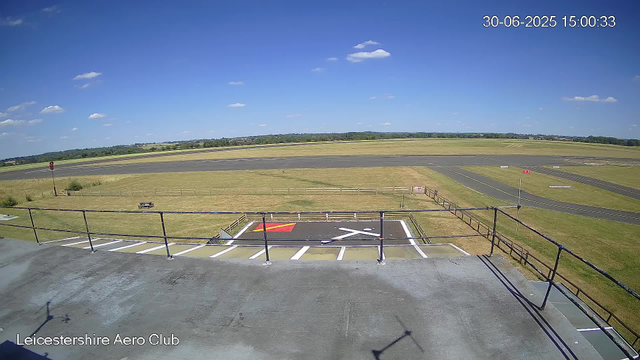A wide view of an airfield with a clear blue sky and scattered small clouds. There is a grassy area with a picnic bench and a small fence, a paved runway in the background, and a marked helipad in the foreground with a white helicopter symbol and red-yellow markings. The image has a timestamp in the upper right corner showing "30-06-2025 15:00:33," and the bottom left corner has the text "Leicestershire Aero Club."