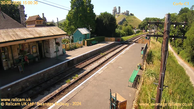 A small train station platform with a building on the left side, benches, and a green trash bin. Railroad tracks run through the center, extending into the distance. To the right, there are wooden utility poles with electrical wires. The area is surrounded by green trees and bushes, with a pathway running parallel to the tracks on the far right. In the background, a hill topped with ancient stone ruins is visible under a clear blue sky.