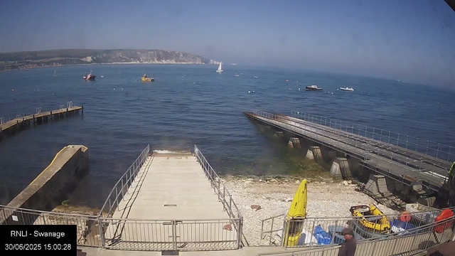 A view of the seaside at Swansea, showing a small pier with metal railings leading into the water, a concrete platform, and a gravel area with yellow safety equipment and supplies. Several boats are anchored in the calm blue sea, with a large white sailboat in the background. The sky is clear with no clouds. Hills or cliffs are visible in the distance across the water. The bottom left corner has a timestamp indicating the date and time.