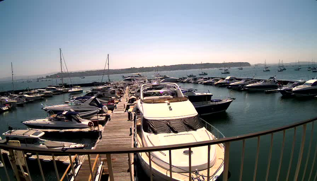 A marina with numerous boats and yachts anchored along wooden docks and in open water. The water is calm, and a distant shoreline with landforms can be seen under a clear sky. The photo appears to be taken from a balcony or a higher vantage point overlooking the marina.