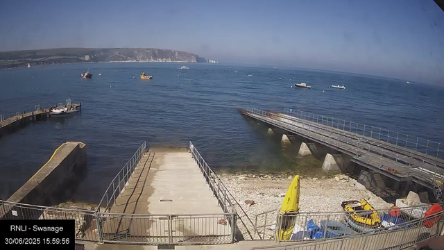 A view of a seaside harbor with a pier extending into the water. Several small boats are floating on the calm sea, and a few are docked near the pier. In the background, there are white chalk cliffs along the coast under a clear blue sky. The foreground shows a small area with equipment and a yellow weather warning cone. The timestamp indicates the date as June 30, 2025, and the time as 15:59:56.