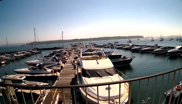 A marina filled with numerous boats and yachts docked along a wooden pier. The water is calm, and the sky is clear with a hint of the setting or rising sun, casting a gentle glow over the scene. In the background, there is a distant shoreline with some hills or land formations. The image is viewed from a vantage point behind a metal railing.