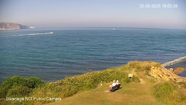 A peaceful seaside scene with two people sitting on a bench near the grass and bushes, facing the calm blue ocean. In the distance, a boat is speeding across the water, leaving a trail of white foam behind. The sky is clear with no clouds, and cliffs with buildings are visible on the left side of the horizon. The timestamp in the top right corner shows the date and time as June 30, 2025, at 16:00:26. The bottom left corner indicates the image is from the Swanage NCI Public Camera.