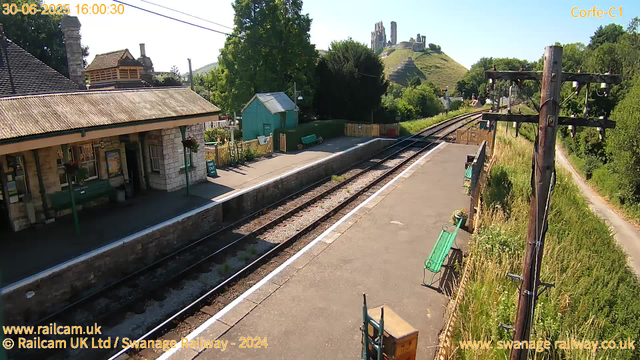 A small train station platform with a building on the left side featuring a roof and stone walls. There are two green benches, a turquoise shed, and a few potted plants. The railway tracks run through the middle, curving into the distance. To the right, there are tall, overgrown grass and trees along a footpath. An old wooden utility pole with electrical wires stands near the edge of the platform, with some maintenance equipment or wires attached. In the background, a hill with ancient ruins or castle remains is visible, and the sky is clear and blue. The scene is bathed in bright sunlight, indicating a sunny day. The date and time shown are June 30, 2025, at 16:00:30.