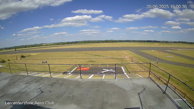 A view from a webcam at Leicesterhire Aero Club showing a clear day with a blue sky and scattered white clouds. In the foreground, there is a small observation or parking area with designated parking spots and a helipad marked with a white "H" and a red square. A bench is situated on the grass near the fence, and a windsock is visible on a pole. In the background, the flat landscape extends to the horizon with grassy fields and a few structures, and there are taxiways and runways on the airfield. The timestamp indicates the date and time as 30th June 2025 at 16:00:36.