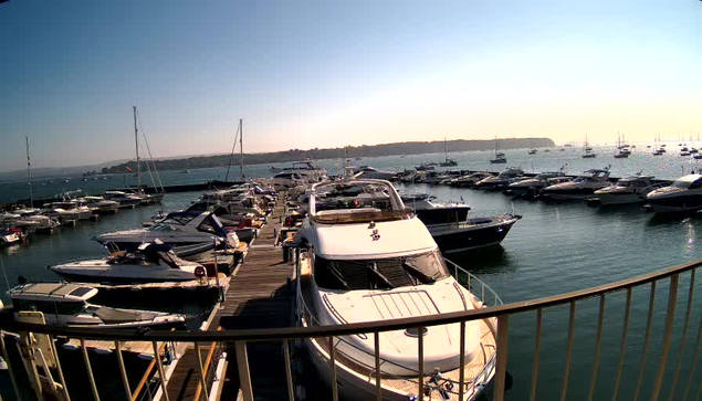 A marina filled with numerous boats and yachts docked along wooden piers, with calm water reflecting the clear sky. In the background, there is a view of a distant shoreline with some land and trees. The photo appears to be taken in the early evening or late afternoon, with sunlight illuminating the scene. A metal railing curves around the foreground.